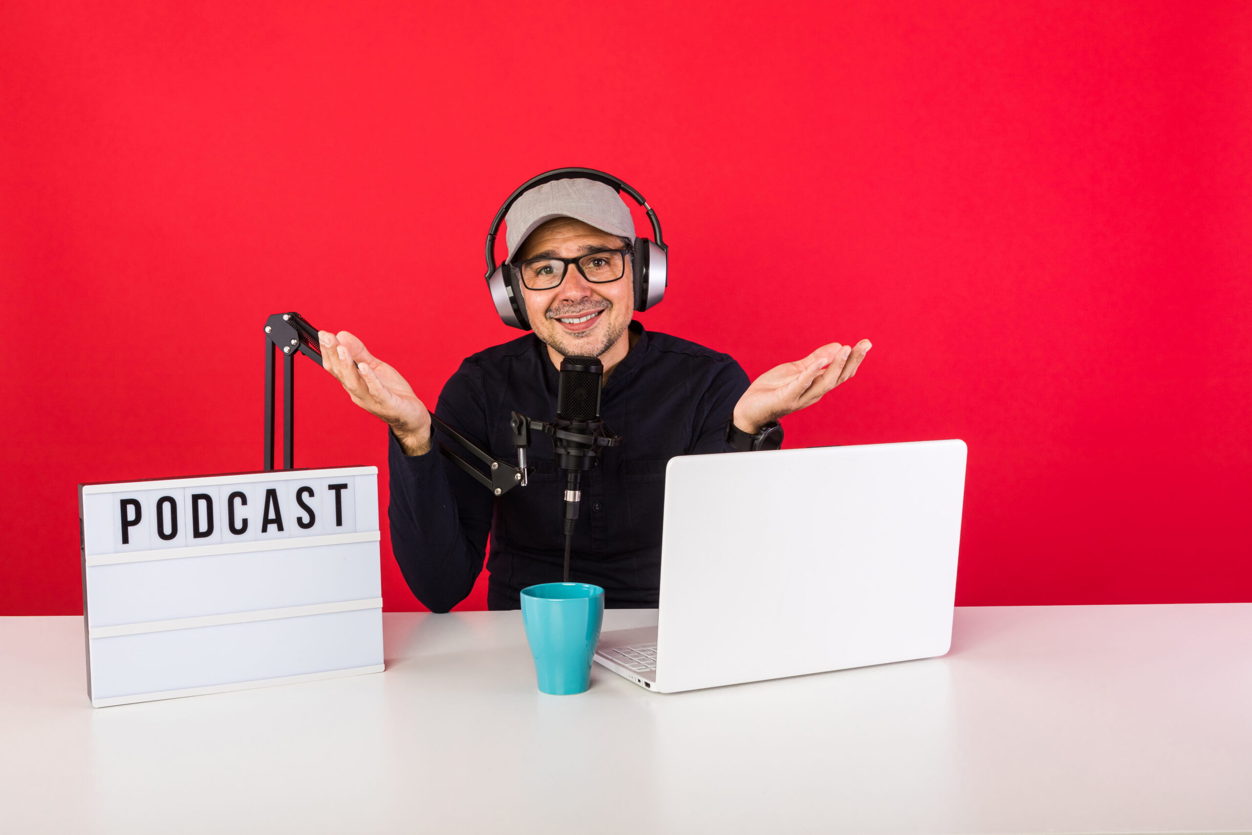 Man presenter with cap in podcast radio recording studio making hand gestures, next to a computer, a microphone and a light box with the word podcast, on red background. Podcasting, broadcast concept